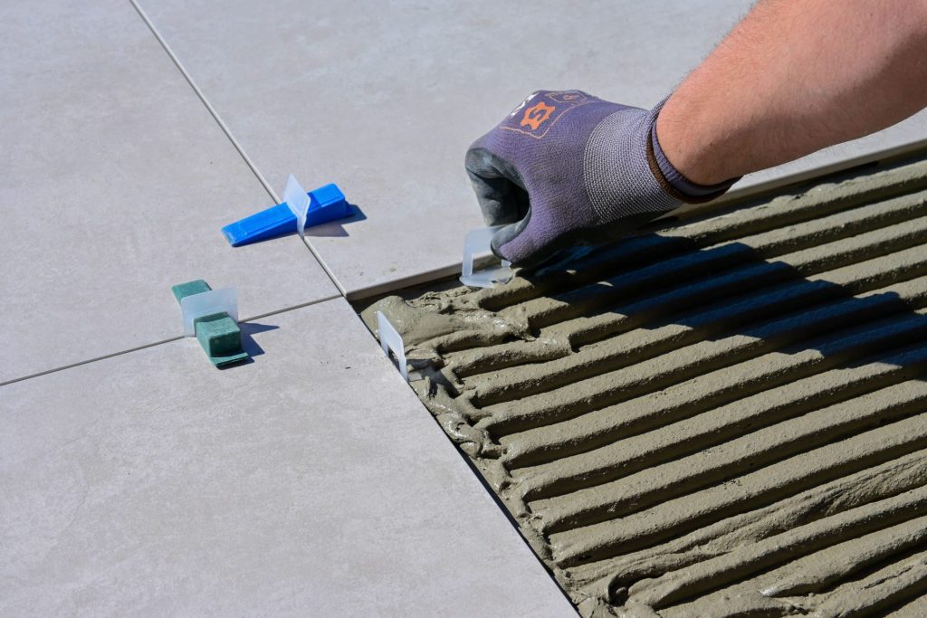 Detailansicht einer behandschuhten Hand, die während der Bauarbeiten Fliesen mithilfe von Nivelliersystemen und Abstandhaltern ausrichtet.