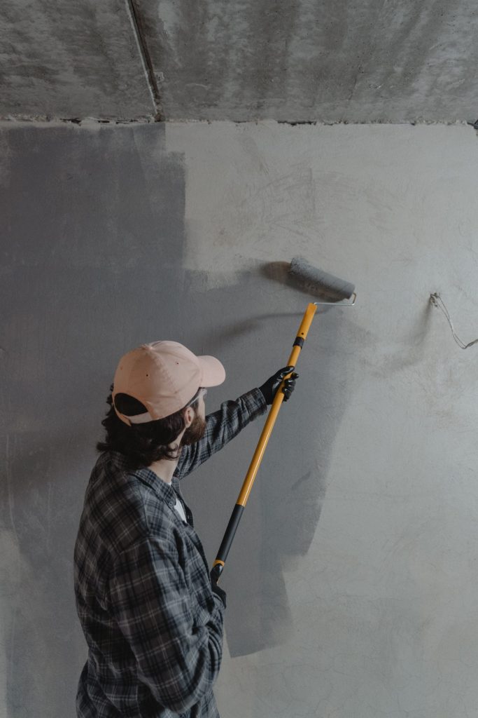 Ein Mann trägt mit einer Farbrolle graue Farbe auf eine Wand in einem unfertigen Raum auf.